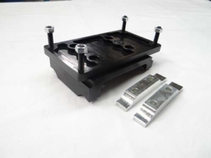 A black metal fixture, identified as the 15" Motor Mounts with a 1/2" standard plate from International Clamps, featuring four bolts, sits next to two silver metal components on a white surface.