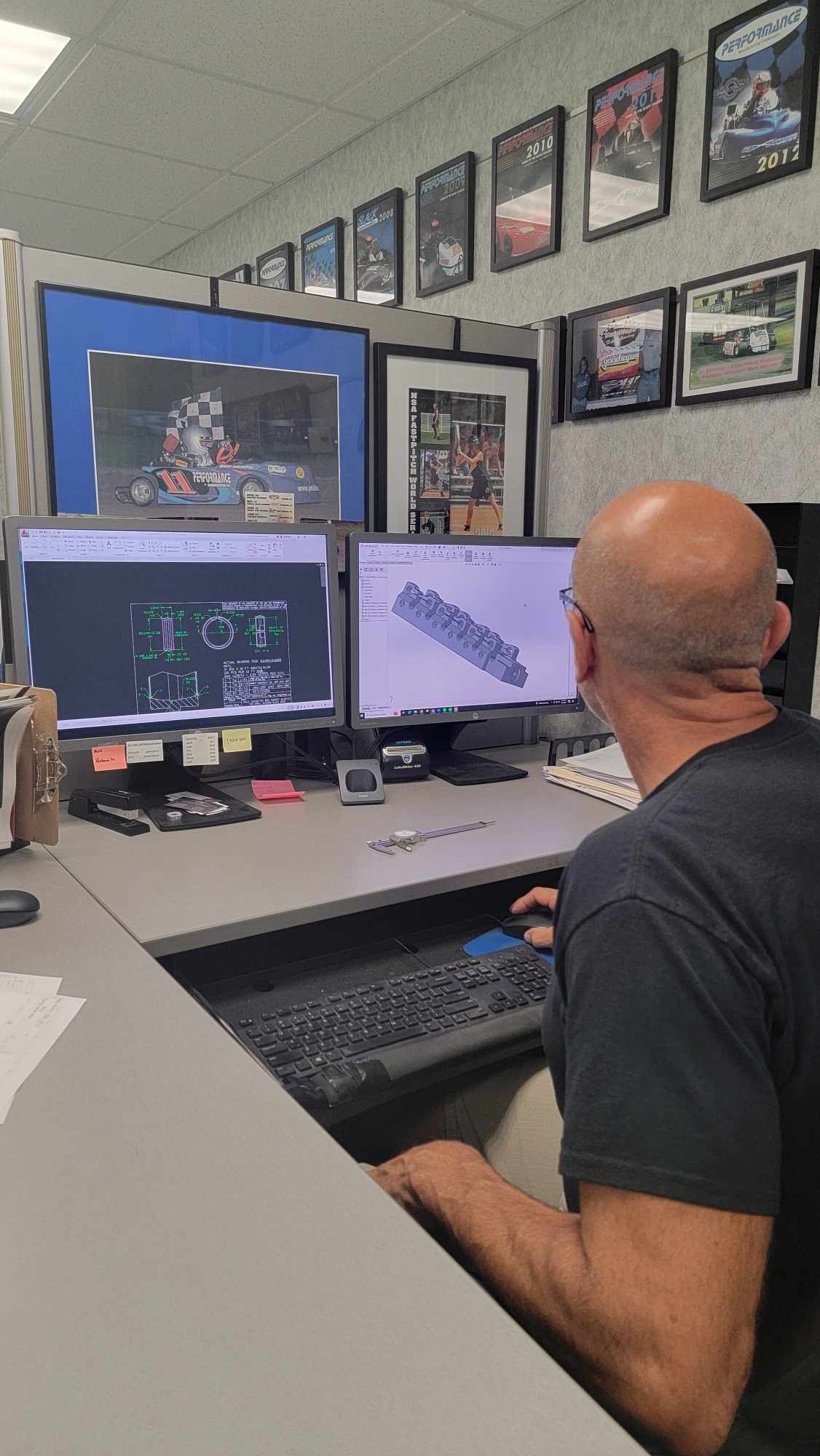 A person is seated at a computer workstation, working on 3D modeling software. Multiple monitors display technical designs. The background shows framed pictures and awards on the wall.