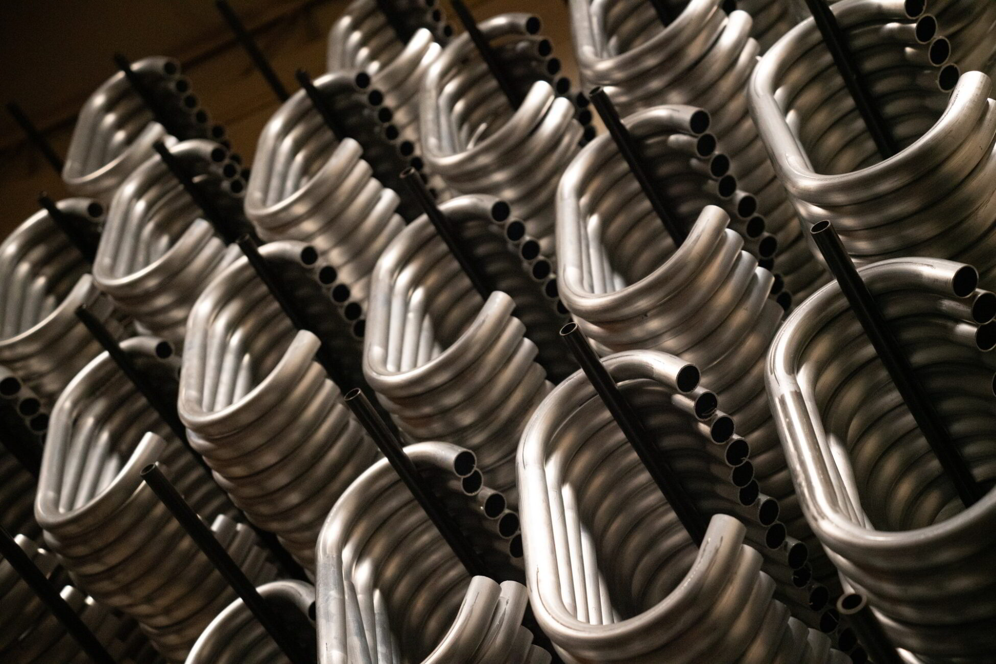 Rows of coiled metal tubes and black rods are stacked vertically in an industrial setting.